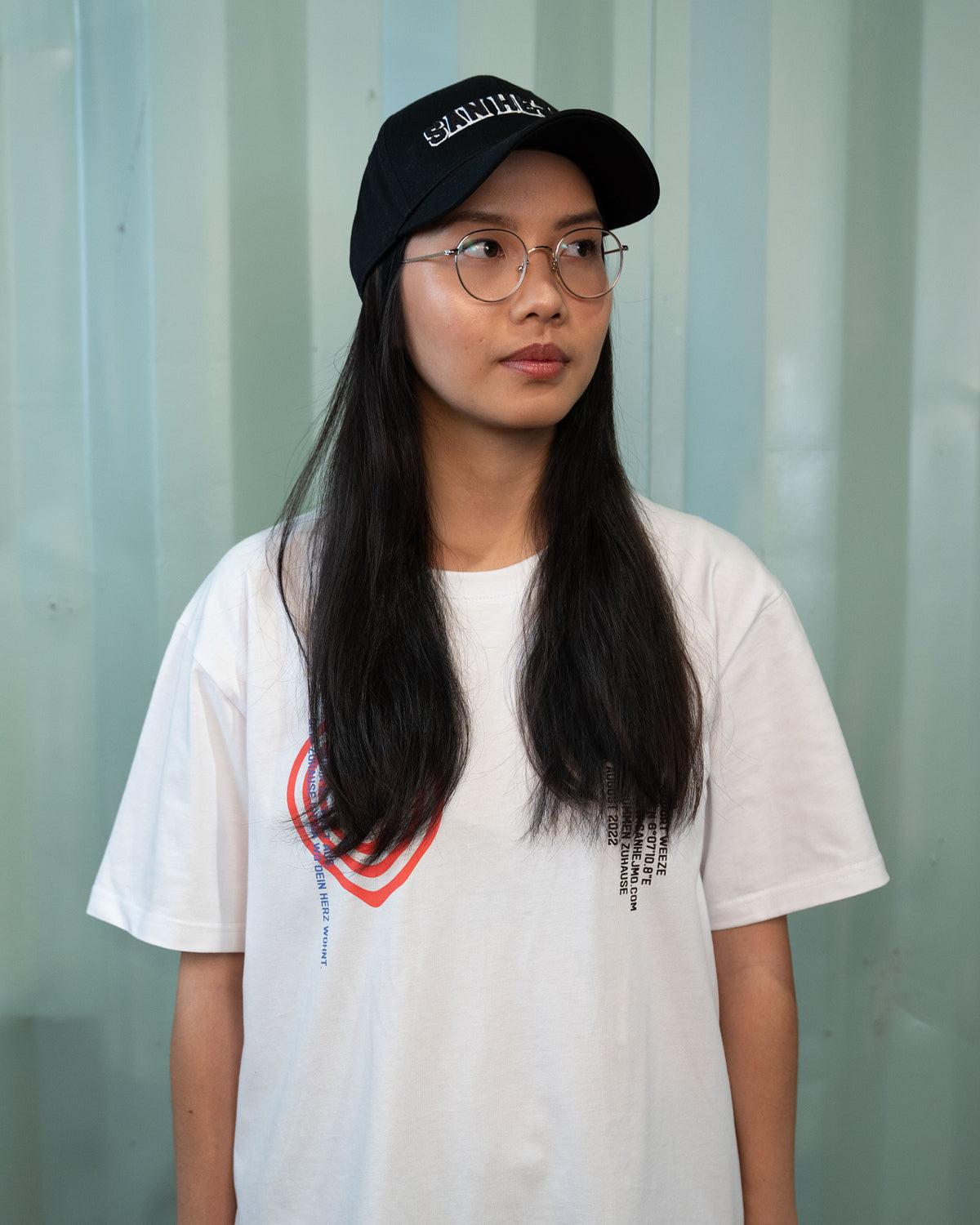 Eine Frau trägt das weiße 2022 Line-Up Shirt und die schwarze Basecap von San Hejmo. Sie guckt zum rechten Bildrand.