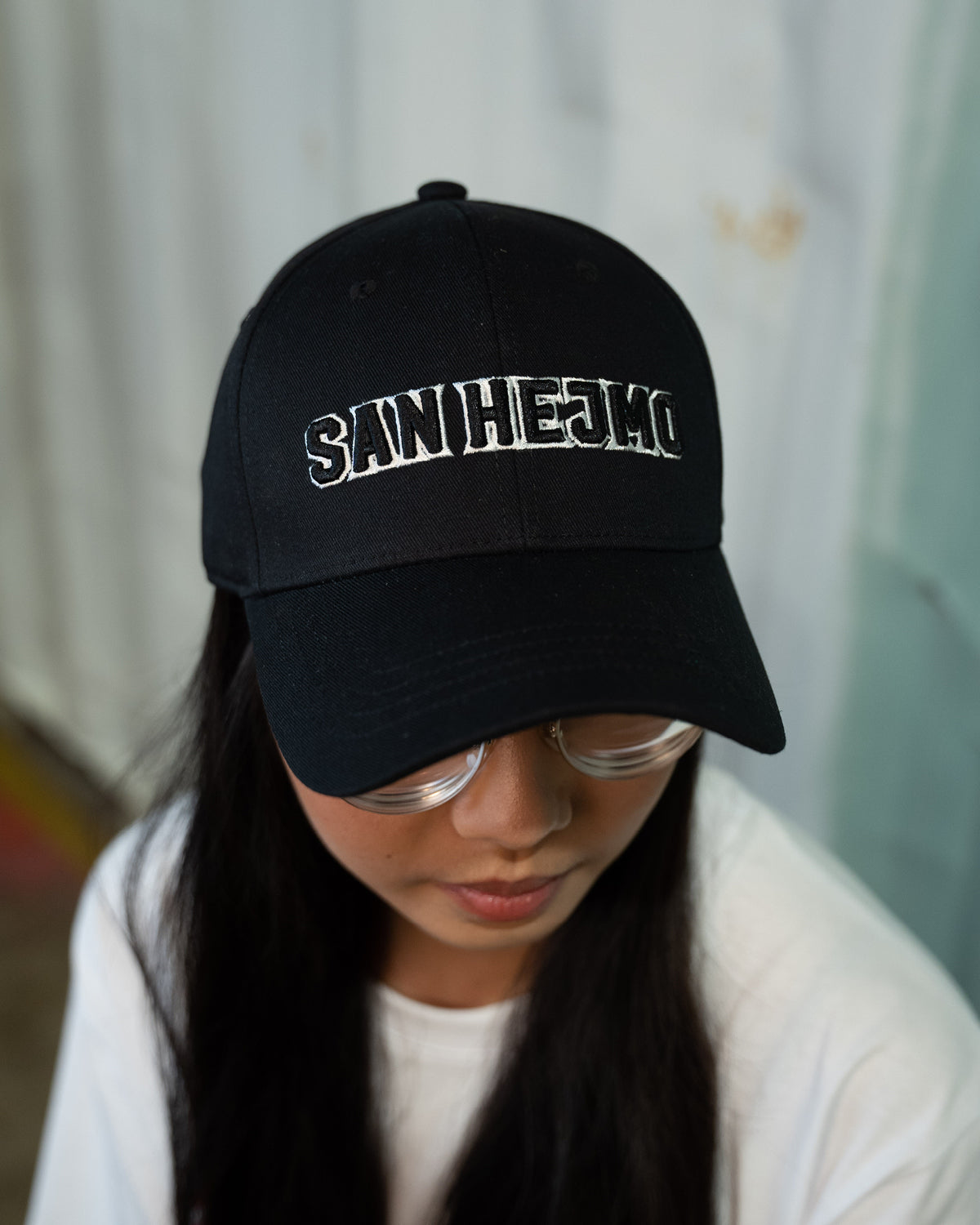 Eine junge Frau trägt die schwarze San Hejmo Basecap und guckt zum unteren rechten Bildrand, so dass die Cap aus der Vogelperspektive zu sehen ist. Ein großer "San Hejmo" Stick in schwarz mit weißen Rahmen um die einzelnen Buchstaben, sitzt vorne mittig.
