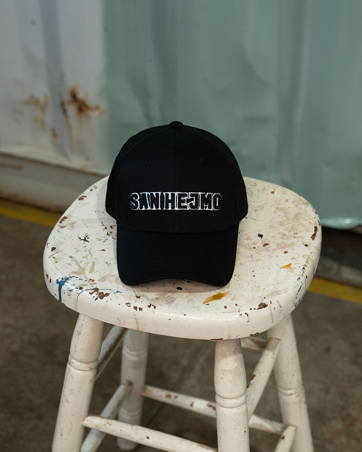 Die San Hejmo Basecap liegt auf einem Hocker, so dass die Cap aus der Vogelperspektive zu sehen ist. Ein großer "San Hejmo" Stick in schwarz mit weißen Rahmen um die einzelnen Buchstaben, sitzt vorne mittig.