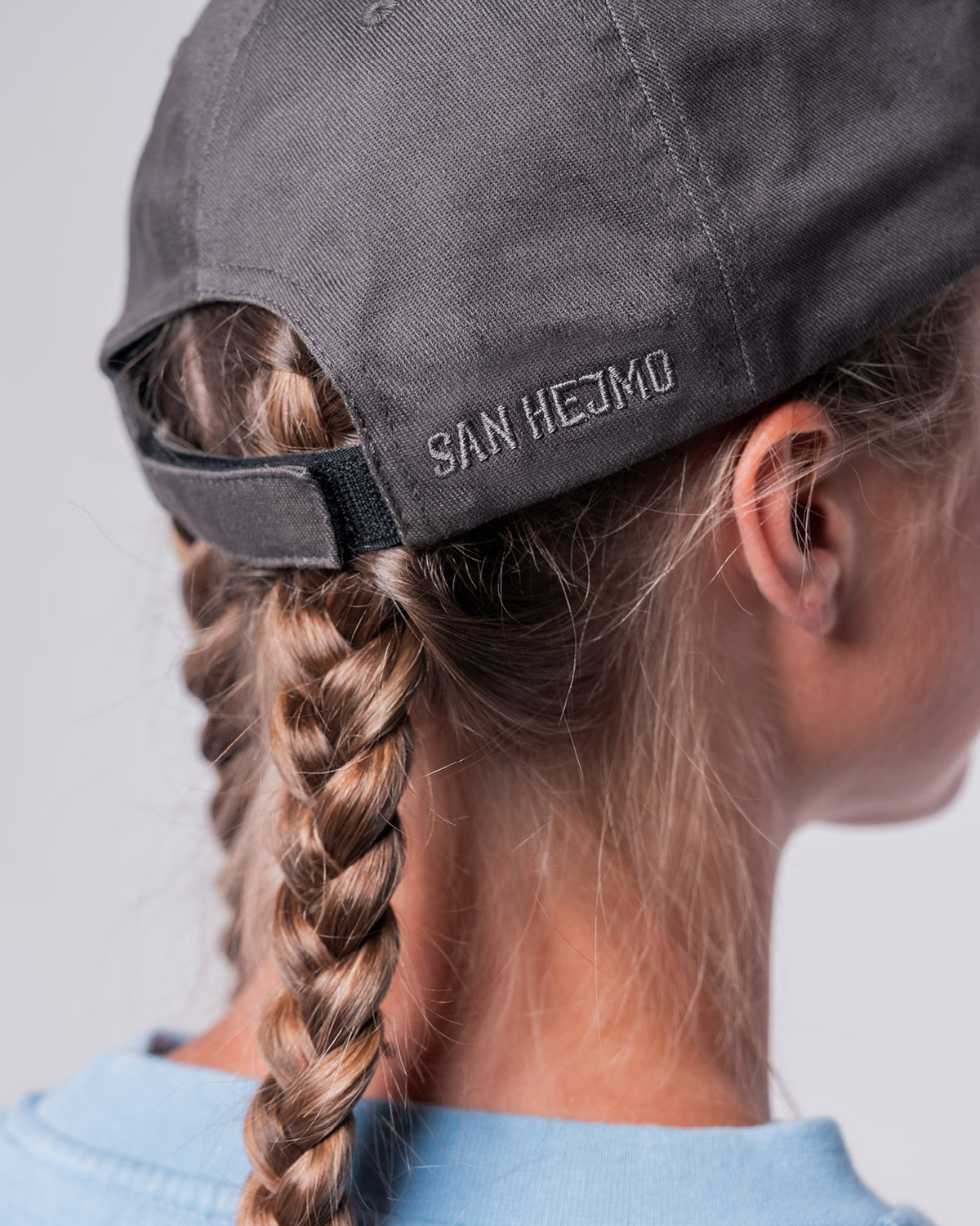 Eine Frau trägt die Anthrazitfarbige Trallafitti Cap. Eine Nahaufnahme der Seite hinten rechts. E sind die Worte "San Hejmo" in schwarz und in Großbuchstaben gestickt. Der Verschluss ist aus Klett.