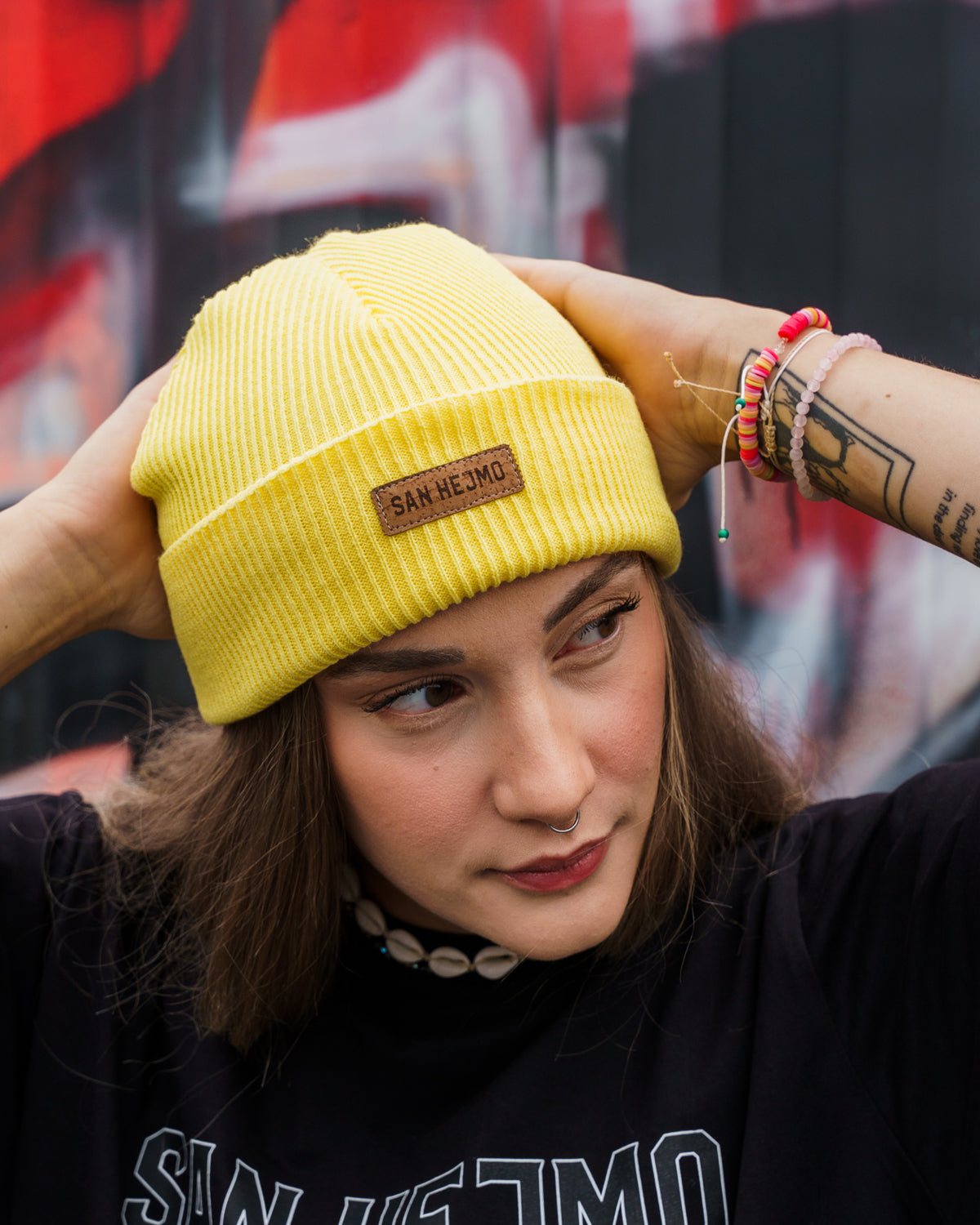Eine Frau vor einer Containerwand, Sie trägt ein schwarzes San Hejmo Oberteil und die gelbe Beanie aus der Basic Collection. Vorne ist ein braunes Tag aufgenäht auf welchem "San Hejmo" in dunkelbraun/schwarz gedruckt ist.