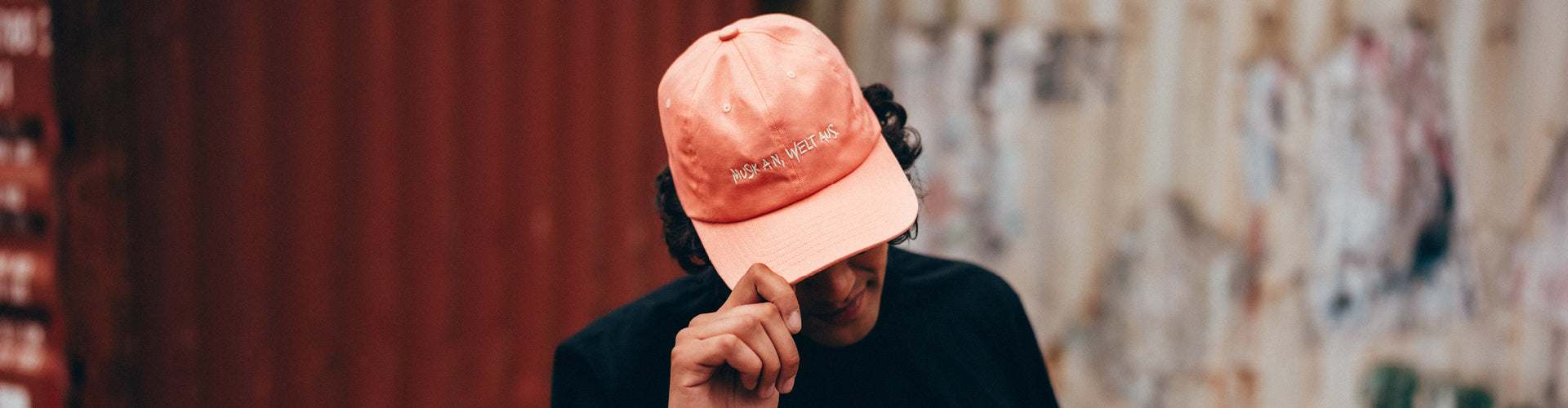 Ein Mann vor einer Containerwand trägt das Line-up T-Shirt und die Apricot farbige Dad Cap. Er hält die Cap mit einer Hand tiefer in sein Gesicht.