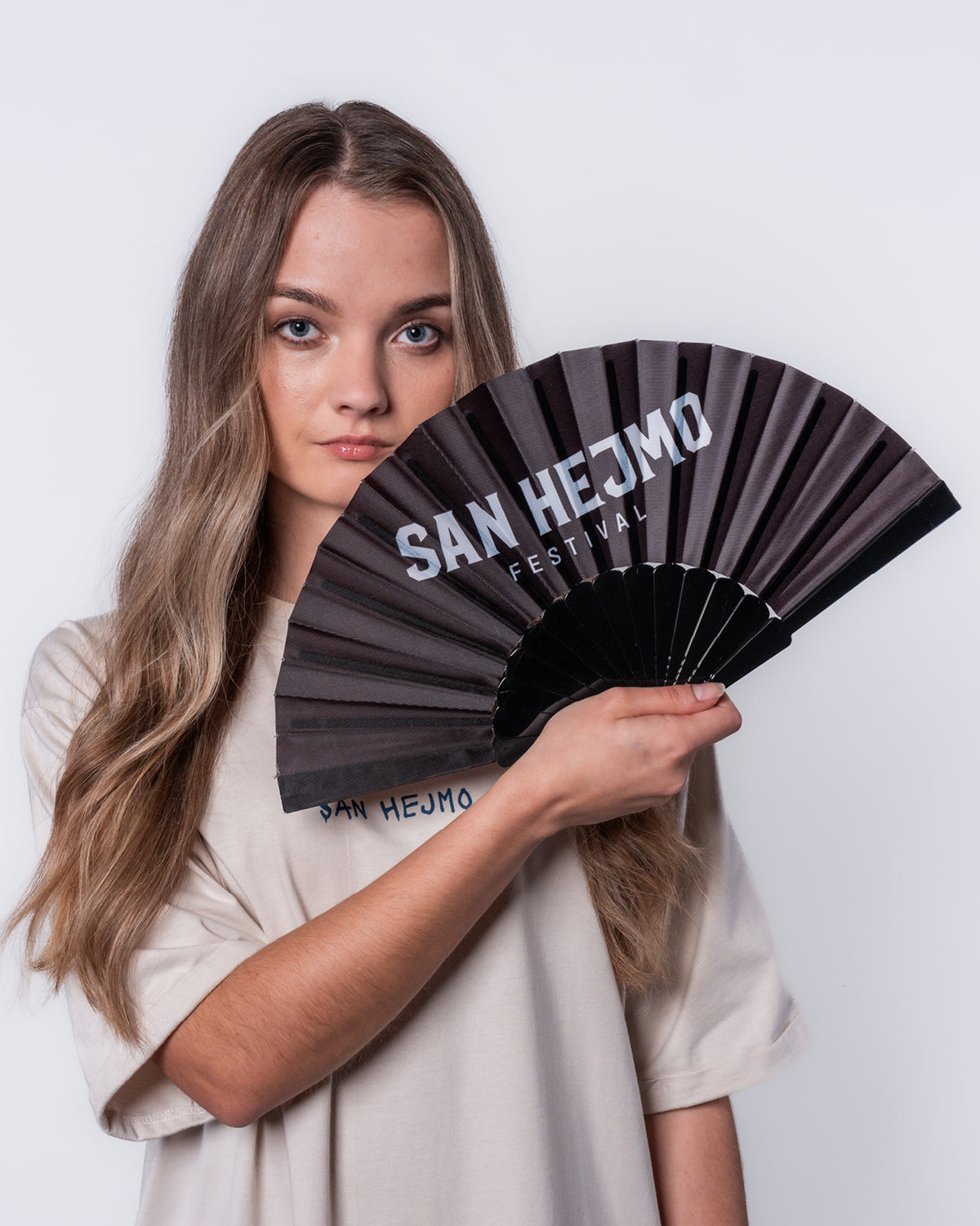 Ein schwarzer Fächer. Auf der oberen Stoffpartie sind die Worte „San Hejmo Festival“ in weiß gedruckt. Gehalten wird er von einer jungen Frau.