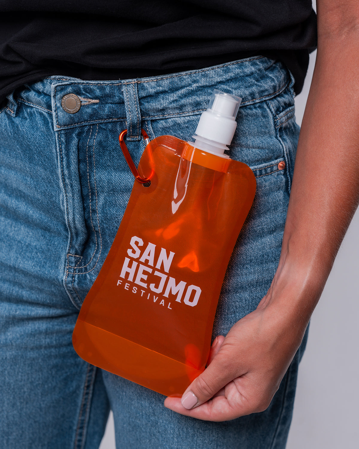 Eine orangene Kunststoffflasche mit einem 450ml Fassungsvermögen und Saugverschluss. Auf der Flasche ist der weiße Aufdruck "San Hejmo Festival" zu sehen. Die Flasche ist mit einem orangenem Karabinerhaken an einer Hose befestigt.