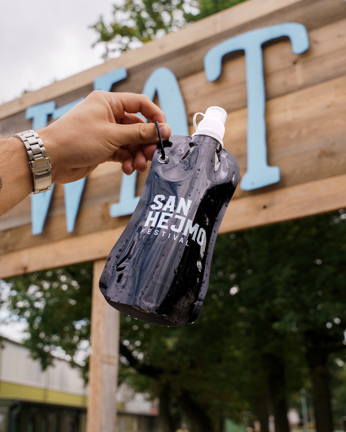 Eine schwarze Kunststoffflasche mit einem 450ml Fassungsvermögen und Saugverschluss. Auf der Flasche ist der weiße Aufdruck "San Hejmo Festival" zu sehen. Die Flasche wird vor eine Wasserstation gehalten.