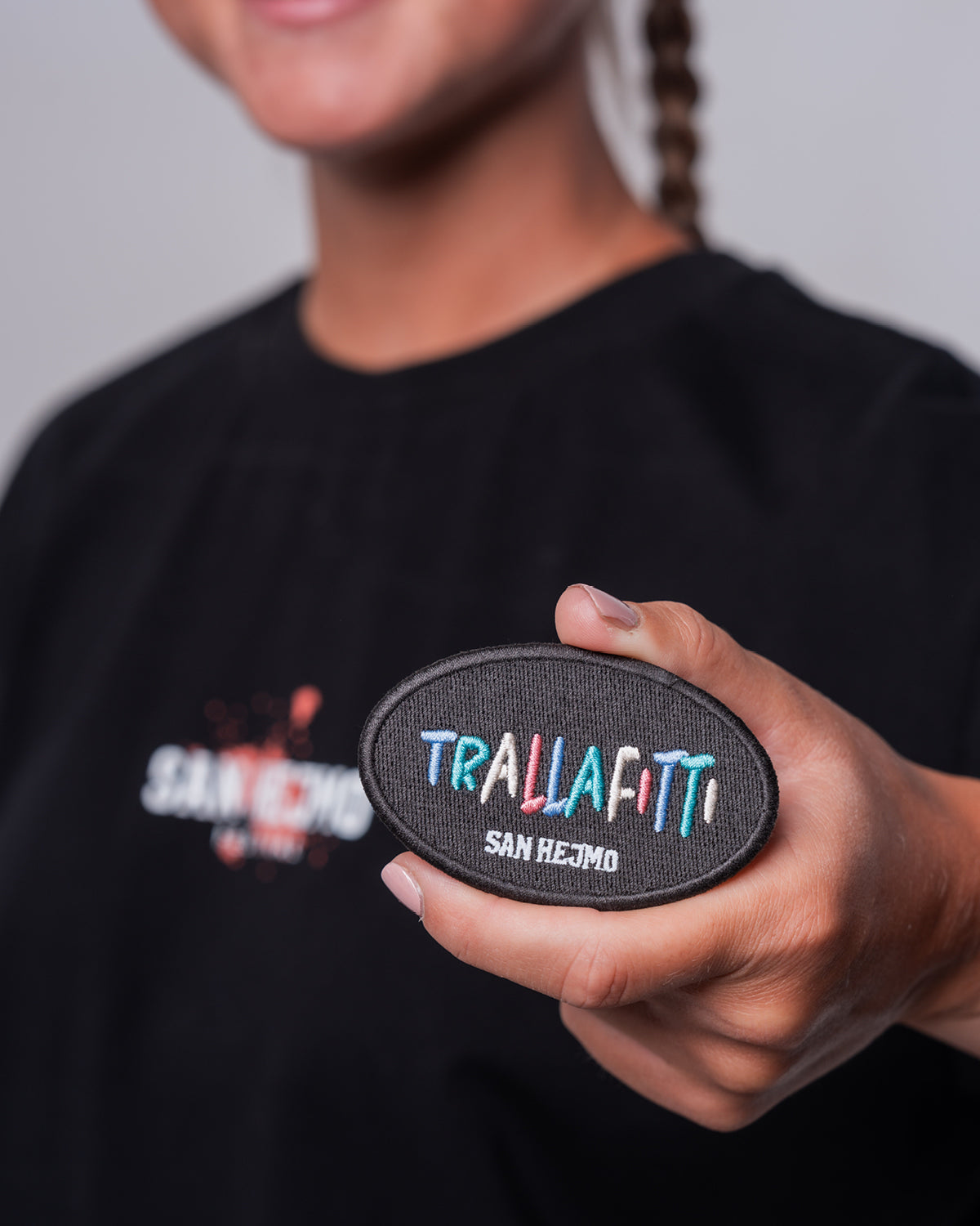 Eine Frau hält einen Magneten, der optisch aussieht wie ein Patch. In jeweils einzeln gestickten bunten Farben steht dort in Großbuchstaben "Trallafitti". Die Schriftgröße ist innerhalb der Buchstaben unterschiedlich. Unter "Trallafitti" steht ebenfalls in Großbuchstaben "San Hejmo" in weiß gestickt.