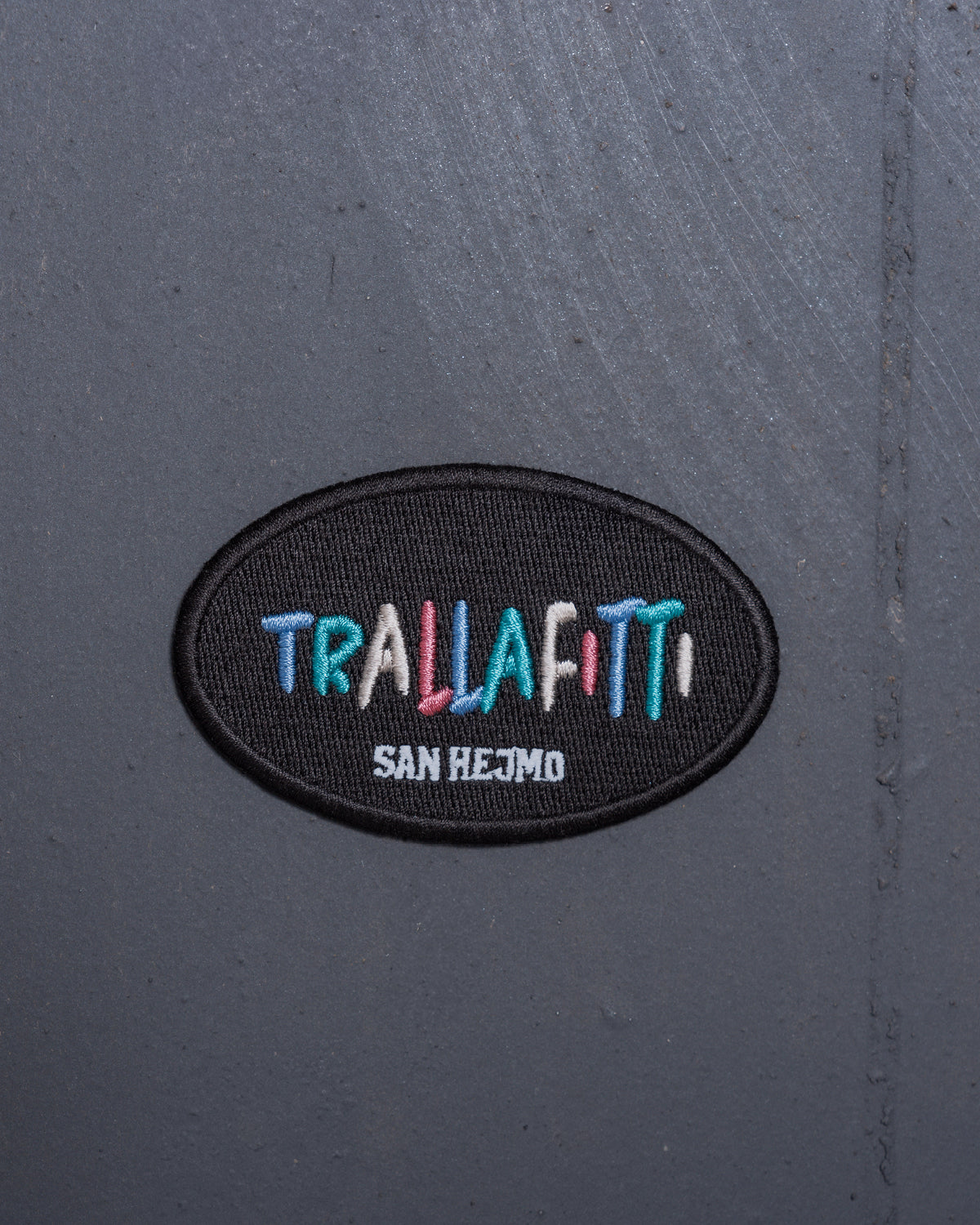 Ein Magnet der optisch aussieht wie ein Patch liegt auf einer grauen Metallwand. In jeweils einzeln gestickten bunten Farben steht dort in Großbuchstaben "Trallafitti". Die Schriftgröße ist innerhalb der Buchstaben unterschiedlich. Unter "Trallafitti" steht ebenfalls in Großbuchstaben "San Hejmo" in weiß gestickt.