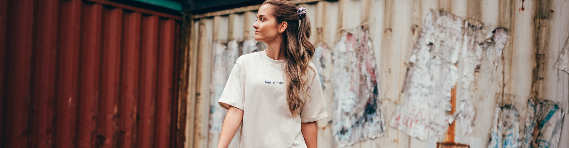 Eine junge Frau steht vor einer Containerwand mit dem Rücken zur Kamera. Sie trägt ein weißes T-Shirt Kleid mit einer einfachen Schrift vorne auf Brusthöhe. Darauf steht in Großbuchstaben "San Hejmo" in dunkler Schrift.