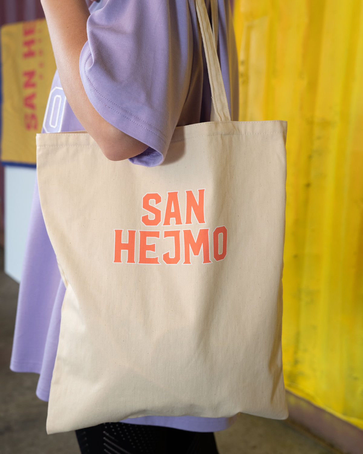 Eine Person trägt einen naturbelassenen, beigen Beutel und ist seitlich gedreht,
damit der Beutel gerade zur Kamera fällt. Mittig auf dem Beutel ist der Schriftzug "San Hejmo" in orange mit kleinem weißen Rahmen um jeden Buchstaben gedruckt.