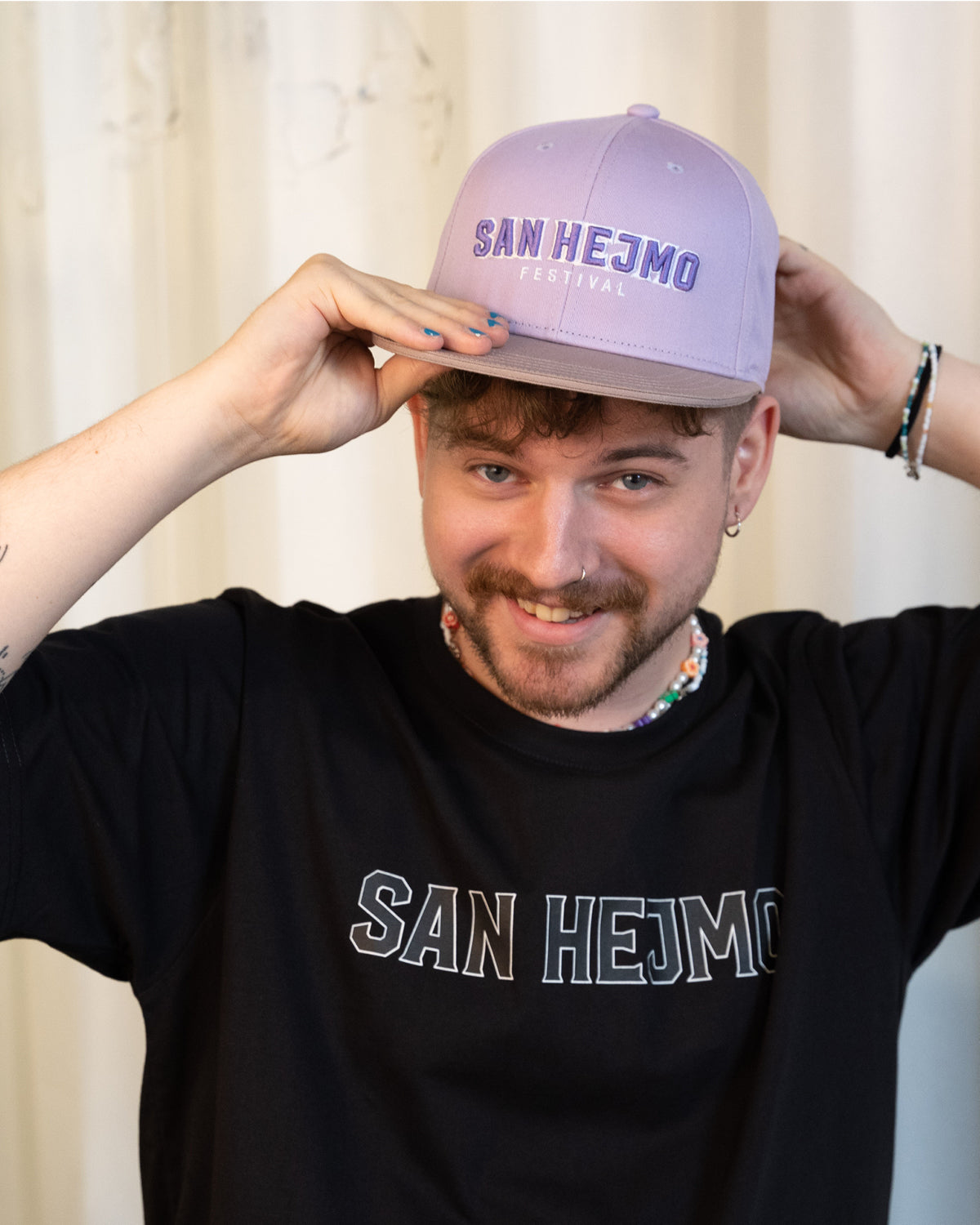 Ein Mann trägt die Lila San Hejmo Basecap und lächelt in die Kamera. Ein großer "San Hejmo" Stick in Lila mit weißen Rahmen um die einzelnen Buchstaben, sitzt vorne mittig.