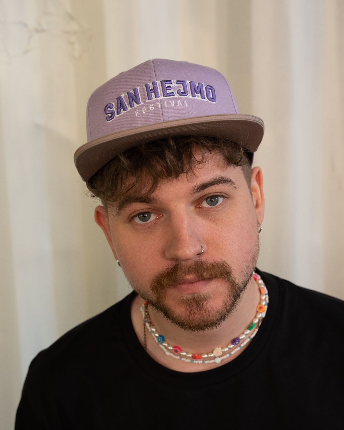 Ein Mann trägt die Lila San Hejmo Basecap und guckt in die Kamera. Ein großer "San Hejmo" Stick in Lila mit weißen Rahmen um die einzelnen Buchstaben sitzt vorne mittig.