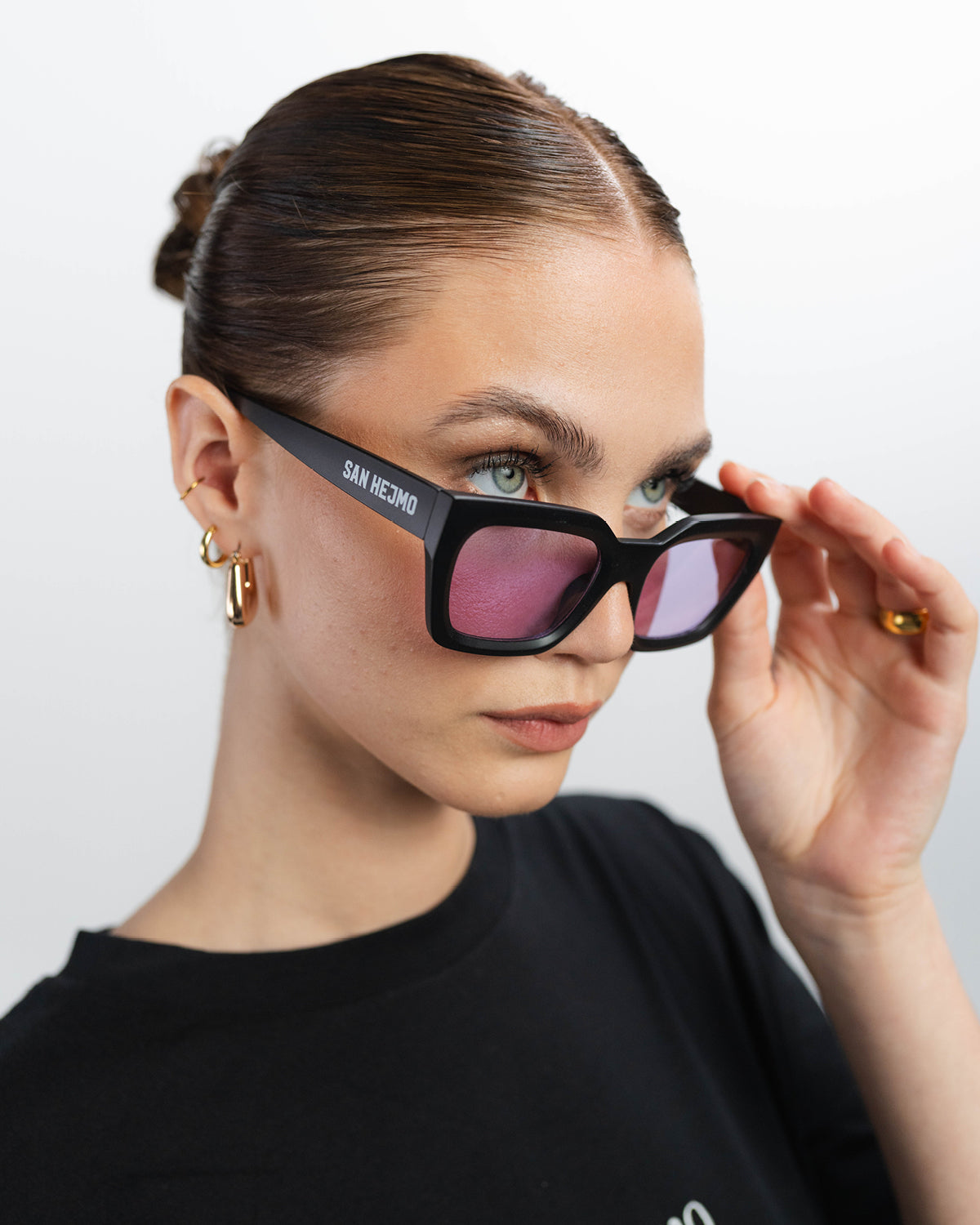 Eine Sonnenbrille mit einem eckig gehaltenen schwarzem Rahmen und rosa Gläsern. Sie wird von einer Frau getragen und schaut über den Rahmen hinaus. Die Brille hat sie dafür ein wenig nach unten geschoben.