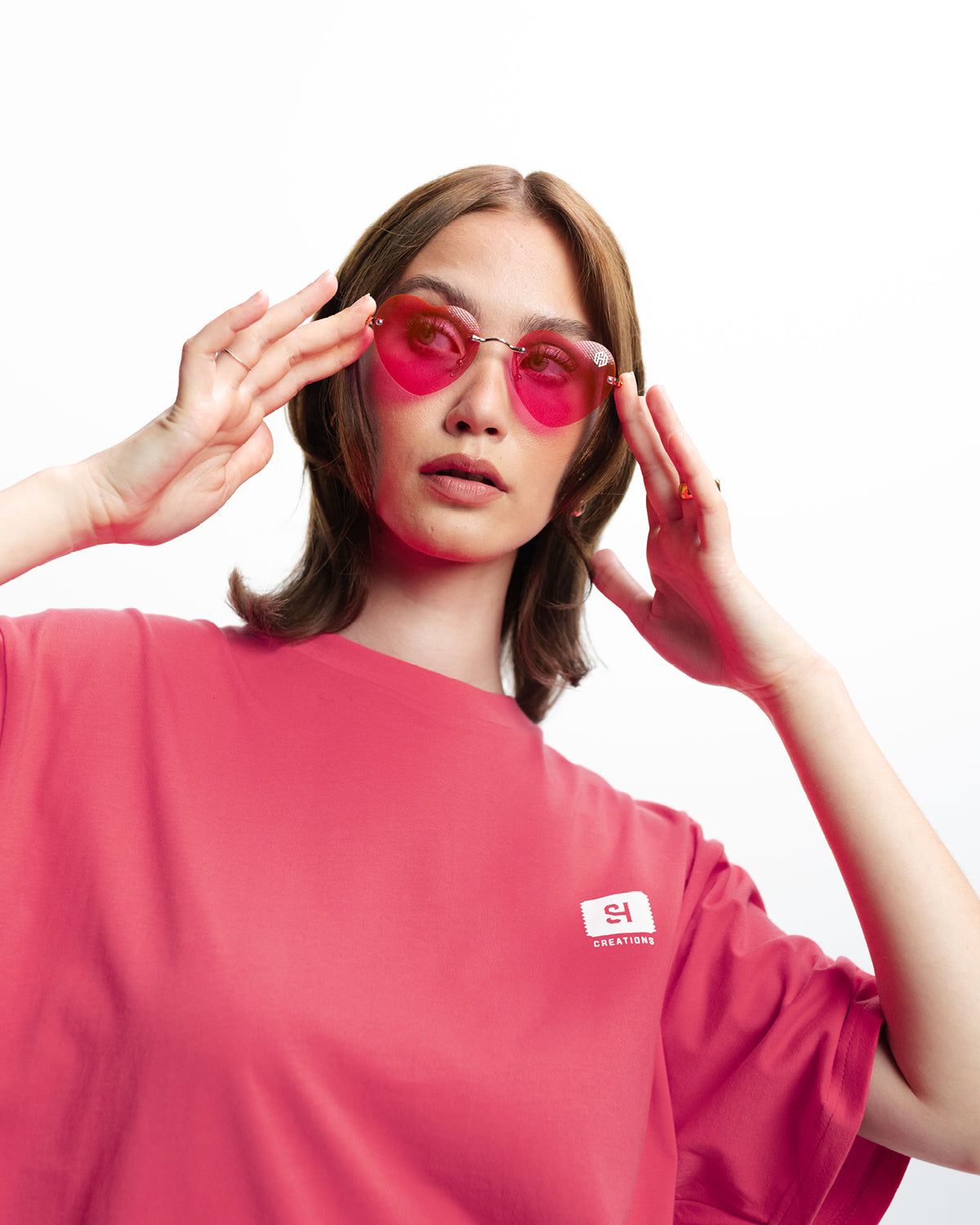 Eine Frau trägt ein pinkes Oberteil und die herzförmige rosa Sonnenbrille von San Hejmo. Sie hält diese rechts und links mit den Fingerspitzen fest.