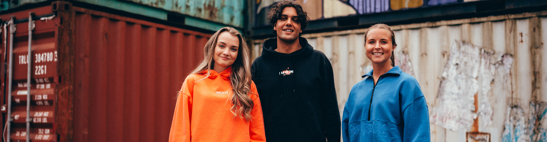 Drei Personen die fröhlich in die Kamera schauen und vor einer Containerwand stehen. Links die Dame trägt einen orangenen Pullover und offene Haare. Der Mann in der Mitte trägt den Schwarzen Line-up Hoodie 2025. Die Frau auf der rechten Seite trägt einen blauen Pullover.