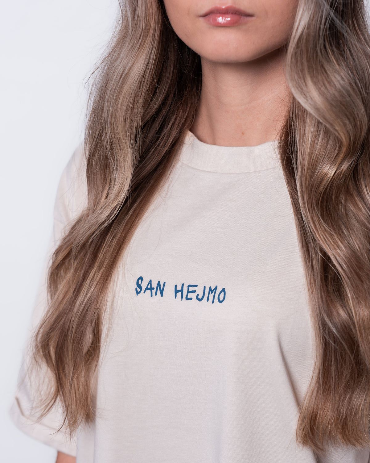 Eine Nahaufnahme von einer jungen Frau mit langen Mittelblonden Haaren. Sie trägt ein weißes Oberteil. Auf Brusthöhe ist in blau "San Hejmo" abgedruckt.