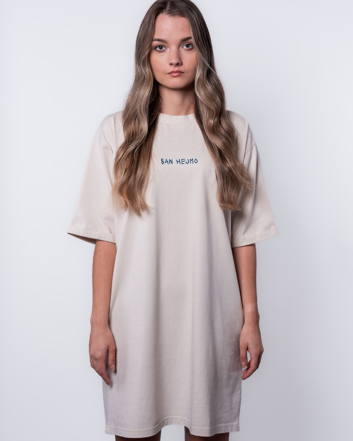 Eine junge Frau steht vor einem weißen Hintergrund, sie trägt ein T-Dress, welches bis über die Oberschenkel geht. Auf der Höhe Ihrer Brust sind die Worte "San Hejmo" in Großbuchstaben gedruckt.