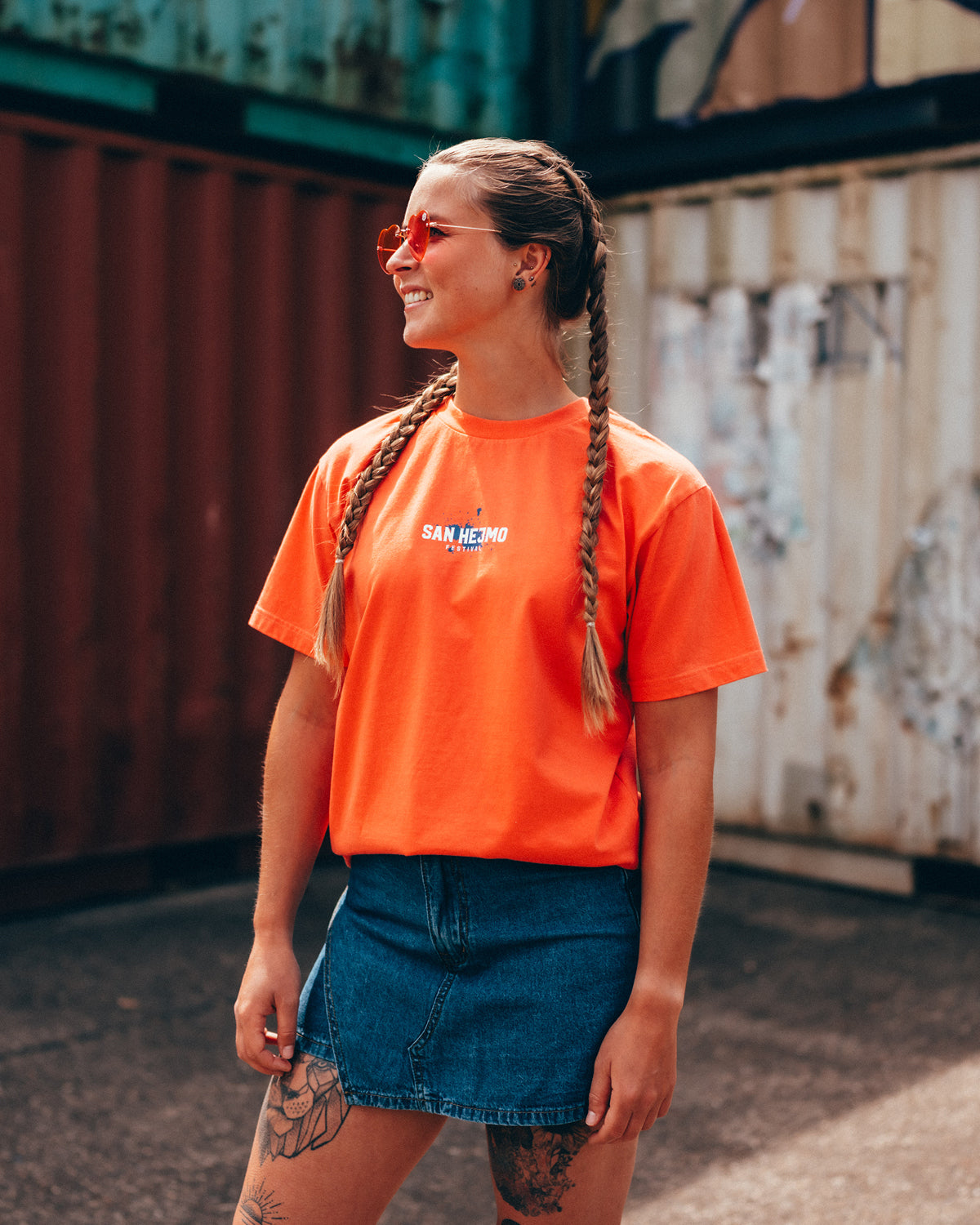 Eine junge Frau steht vor einer Containerwand. Sie hat zwei lange geflochtene Zöpfe, trägt einen kurzen Jeansrock, ein T-Shirt in Coral und eine pinke Sonnenbrille in Herzform. Ihr Blick ist nach links.