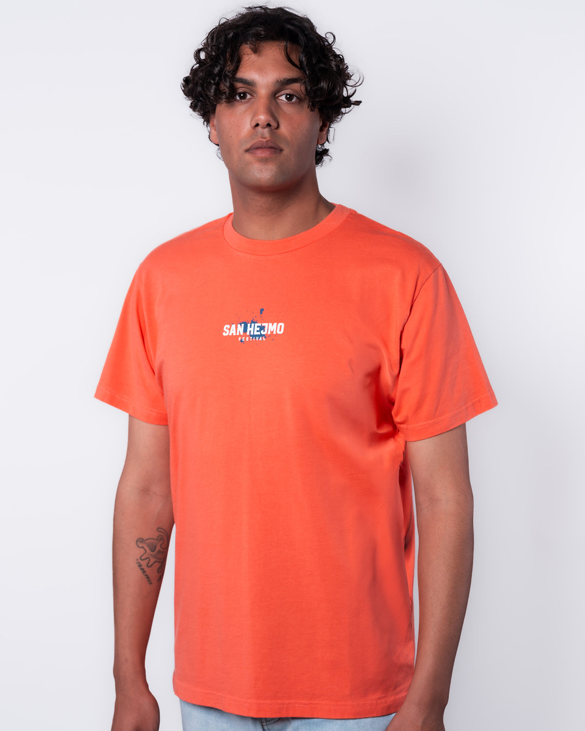 Ein junger Mann mit kurzen schwarzen Haaren steht vor einem weißen Hintergrund und trägt das Coral farbige Line-up Shirt aus 2025. auf Brusthöhe ist in weiß "San Hejmo" und "Festival" zu lesen. Unter der weißen Schrift in ein blauer Farbklecks hinterlegt.