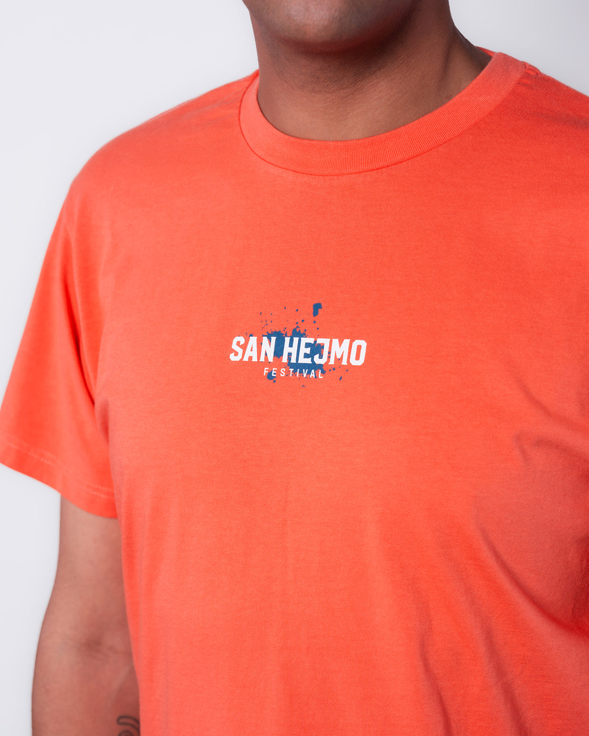 Das Coral farbige Line-up Oberteil 2025 mit weißer Schrift auf Brusthöhe. dort steht "San Hejmo" und in klein darunter "Festival". Die Schrift ist mit einem kleinen blauen Farnklecks hinterlegt.