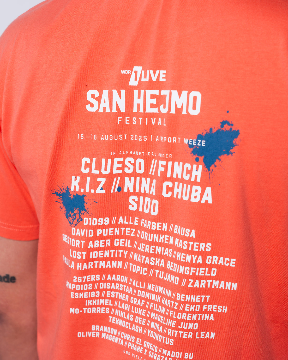 Ein Bild vom Rückenprint des San Hejmo Line-up 2025 T-Shirts in der Farbe Coral. Das Line-up ist in weißer Schrift gedruckt. Oben rechts und mittig links ist hinter der Schrift jeweils ein blauer Klecks gedruckt. Über dem Line-up ist das San Hejmo Logo gedruckt.