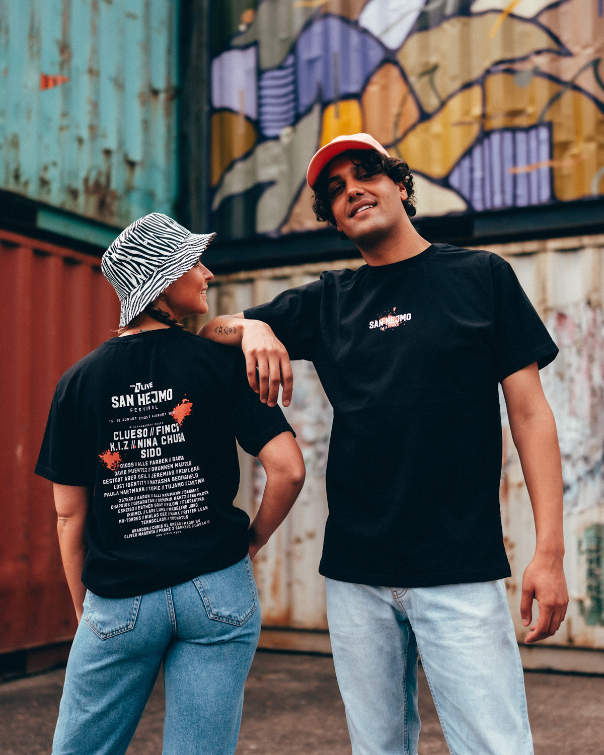 Eine junge Frau und ein junger Mann vor einer Containerwand. Sie tragen beide blaue Jeans und das San Hejmo Line-up 2025 Oberteil in Schwarz. Sie trägt einen Zebra Bucket Hat und er eine Coral farbige Cap.