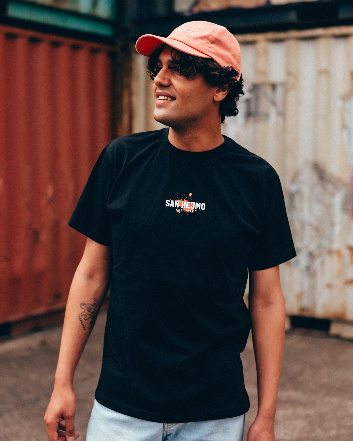 Ein junger Mann mit dunklen Haaren trägt das San Hejmo Line-up 2025 T-Shirt. Auf der Brust ist in weiß die Aufschrift "San Hejmo" und darunter "Festival" gedruckt. Im Hintergrund ist ein kleiner Coral farbiger Klecks.