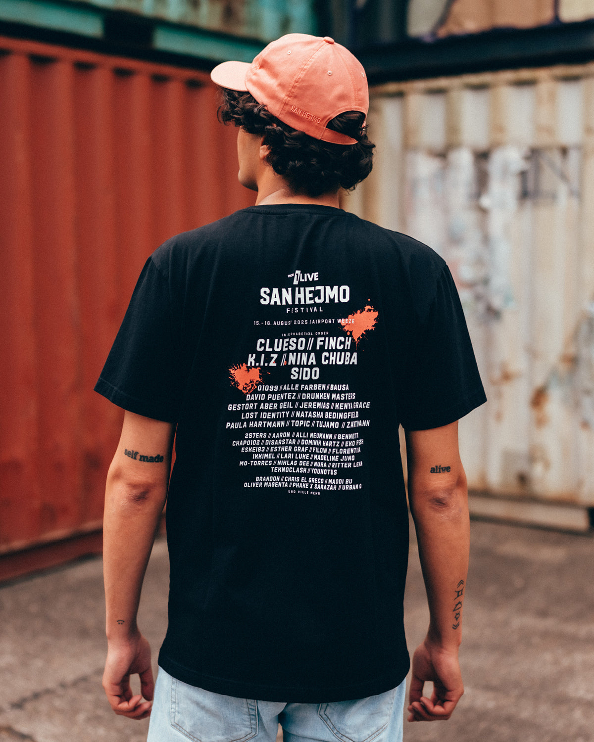 Ein junger Mann steht vor einer Containerwand und trägt das schwarze San Hejmo Line-up Oberteil.
Er steht mit dem Rücken zur Kamera. Der weiße Rückenprint ist oben rechts und mittig links mit jeweils einem Coral farbigen Klecks hinterlegt.