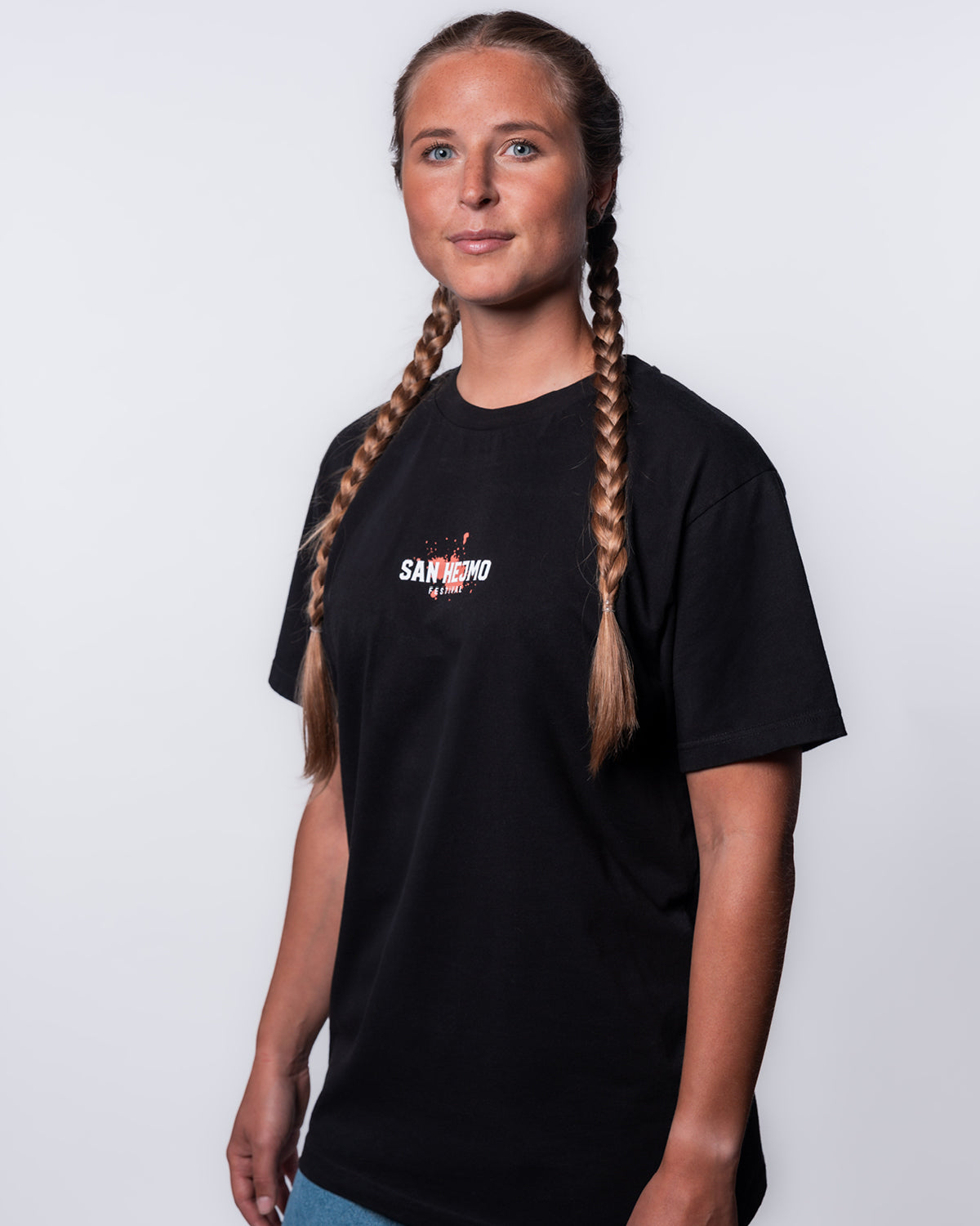 Eine junge Frau mit geflochtenen blonden Haaren steht vor einer weißen Wand. Sie trägt das schwarze San Hejmo Line-up 2025 T-Shirt . Auf Brusthöhe ist in weißen Großbuchstaben "San Hejmo" und eine Zeile darunter und in kleiner Schrift "Festival" gedruckt, Die weiße Schrift in ist meinem Coral farbigen Klecks hinterlegt.