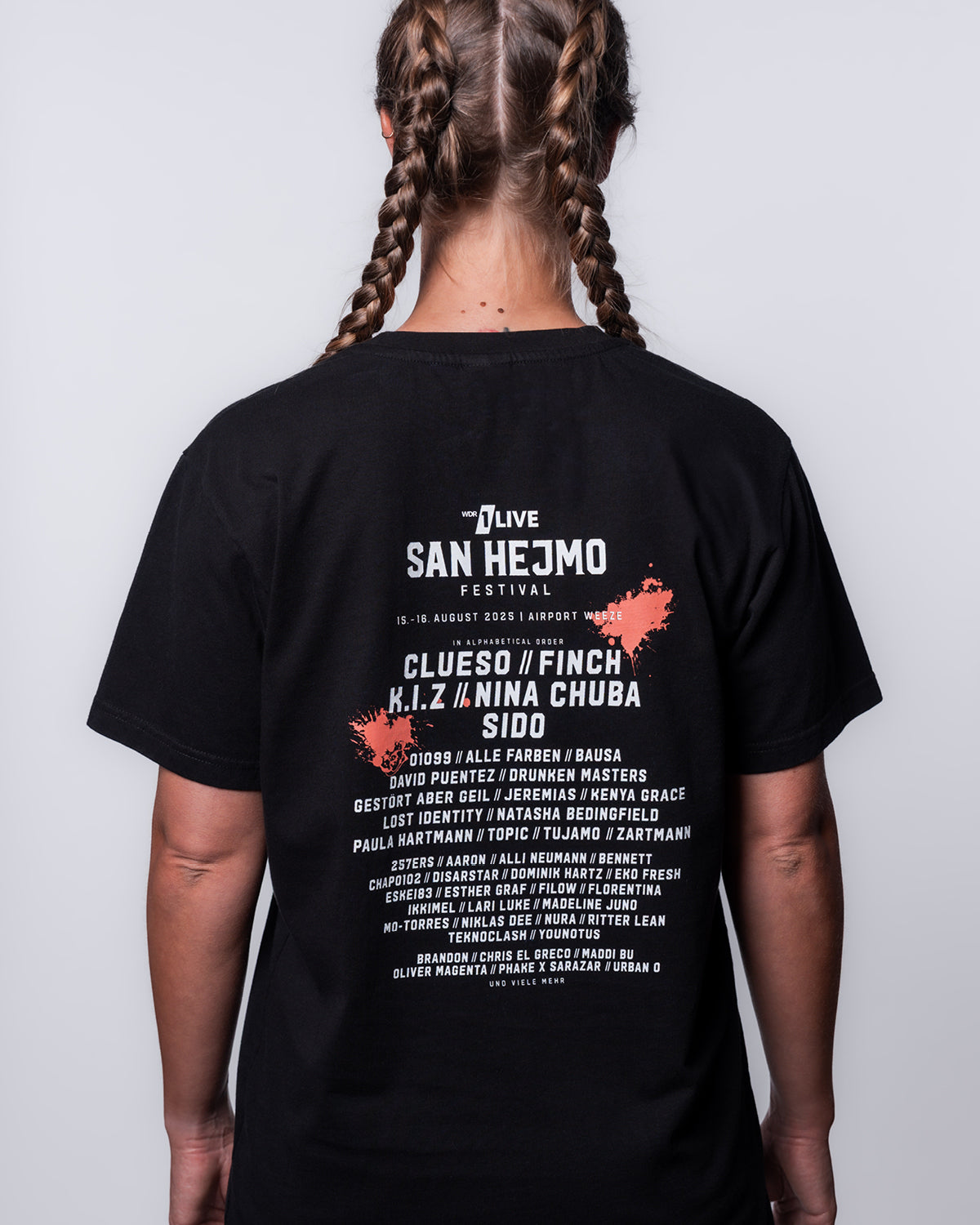 Eine Oberkörper-Aufnahme vom Rückenprint des San Hejmo Line-up 2025 Oberteils in schwarz. Oben ist in weißer Schrift der Name des Festivals gedruckt. Die Headliner sind darunter abgebildet. In kleinerer Schrift finden sich wiederum darunter die weiteren Artists. Oben rechts und unten links ist jeweils ein Coral farbiger Klecks gedruckt.