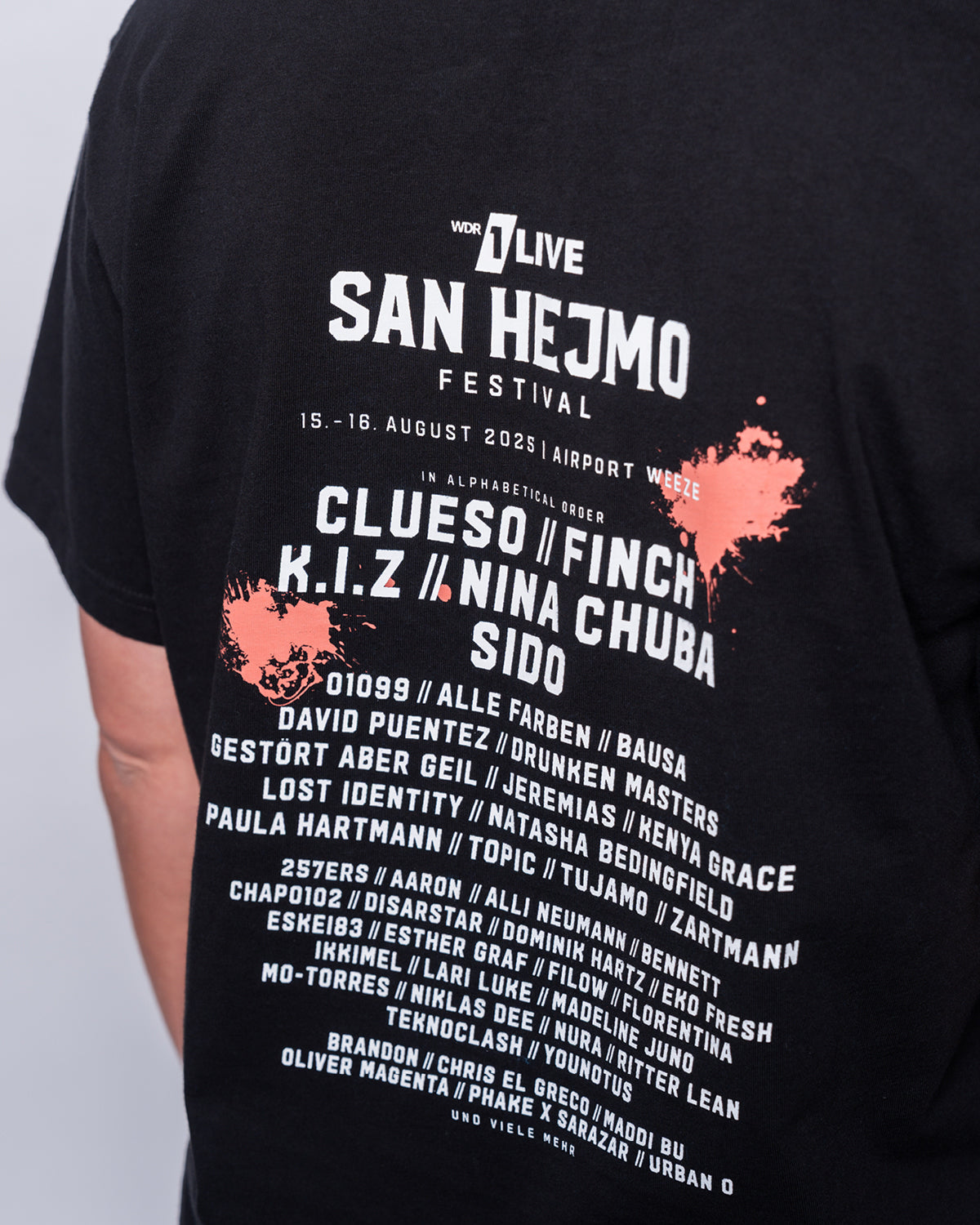 Eine Nahaufnahme vom Rückenprint des San Hejmo Line-up 2025 Oberteils in schwarz. Oben ist in weißer Schrift der Name des Festivals gedruckt. Die Headliner sind darunter abgebildet. In kleinerer Schrift finden sich wiederum darunter die weiteren Artists. Oben rechts und unten links ist jeweils ein Coral farbiger Klecks gedruckt.