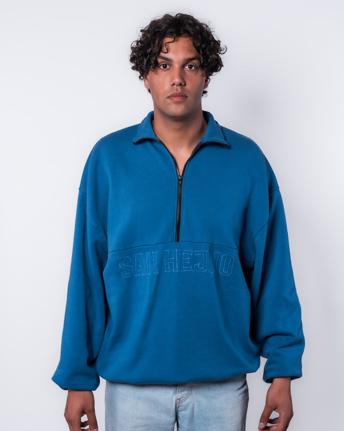 Ein junger Mann trägt den blauen San Hejmo Troyer. Unter der mittigen Längsnaht ist in Großbuchstaben "San Hejmo" gestickt. Der schwarze Half-Zip ist mittig bis zur Längsnaht und geht bis zum Ende des Kragens. Der Kragen des Troyers ist umgeklappt.