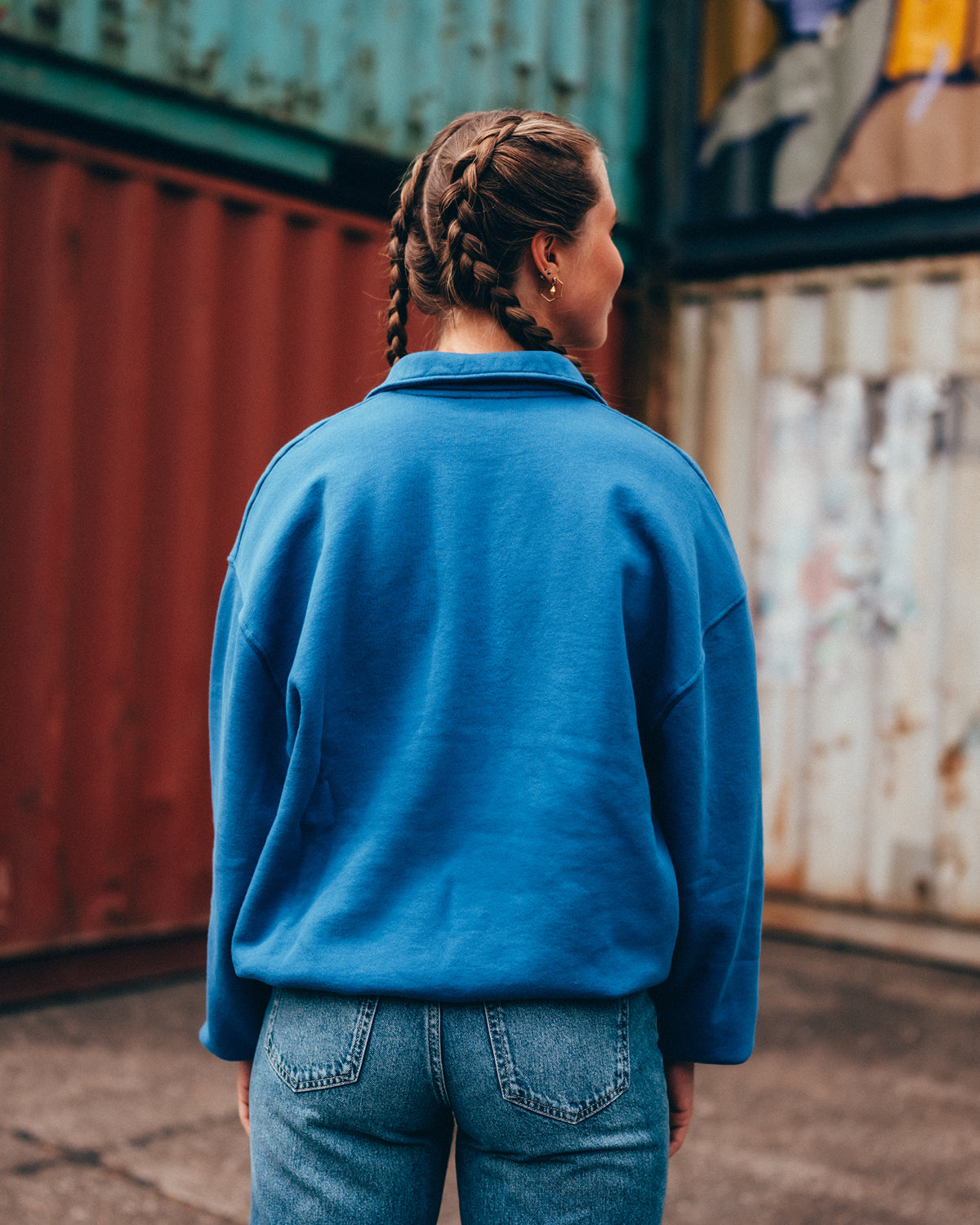 Eine junge Frau steht vor einer Containerwand mit dem Rücken zu Kamera. Sie trägt den blauen San Hejmo Troyer. Der Rücken ist ohne Druck oder Stick.