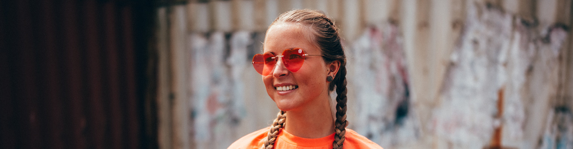 Eine Frau trägt das Coral farbige Line-up Oberteil und die herzförmige Sonnenbrille mit rot-pinken Gläsern. Man sieht ihre Augen durch das Glas. Sie schaut zum linken Bildrand und lächelt.