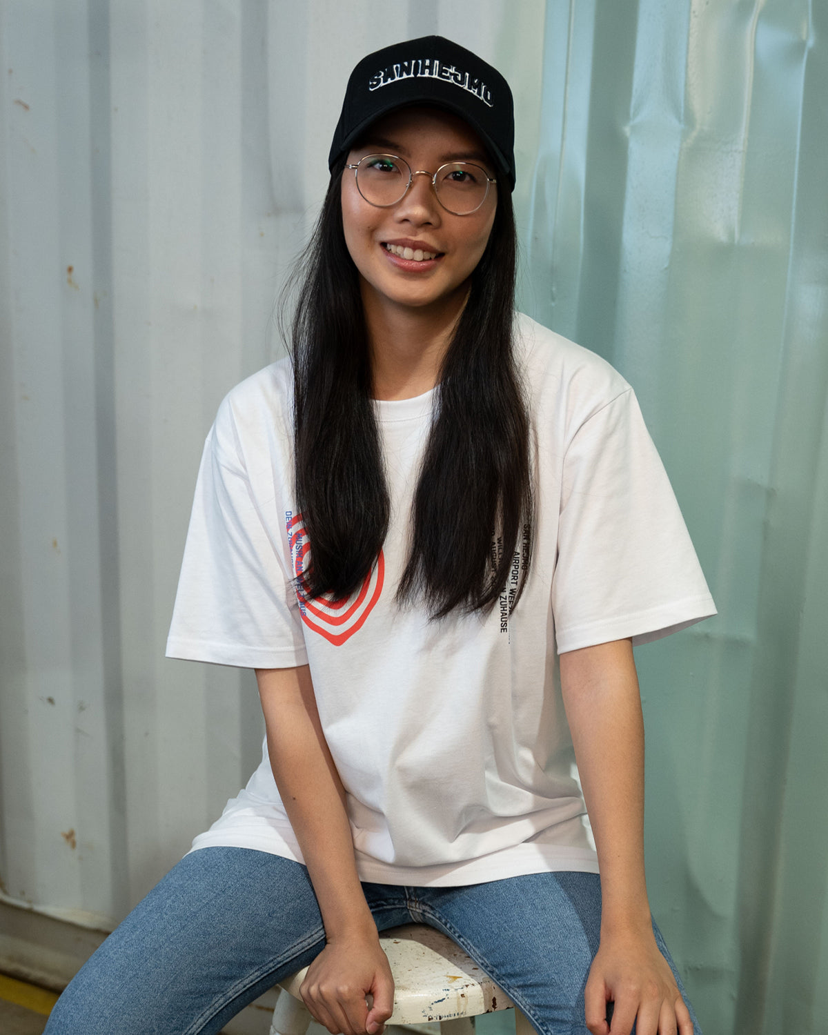 Eine Frau trägt das weiße 2022 Line-Up Shirt und die schwarze Basecap von San Hejmo. Sie lächelt in die Kamera und sitzt auf einem Hocker