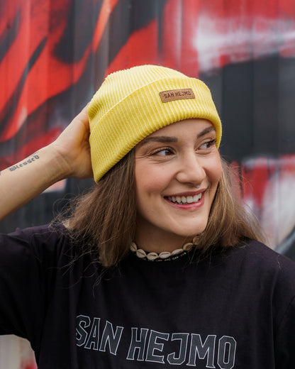 Eine Frau vor einer Containerwand, Sie trägt ein schwarzes San Hejmo Oberteil und die gelbe Beanie aus der Basic Collection. Vorne ist ein braunes Tag aufgenäht auf welchem "San Hejmo" in dunkelbraun/schwarz gedruckt ist.