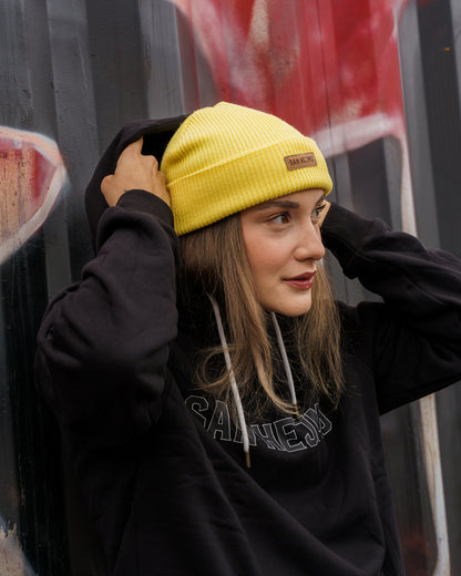 Eine Frau vor einem angesprühten Container, Sie trägt einen schwarzen San Hejmo Pullover und die gelbe Beanie aus der Basic Collection. Vorne ist ein braunes Tag aufgenäht auf welchem "San Hejmo" in dunkelbraun/schwarz gedruckt ist.