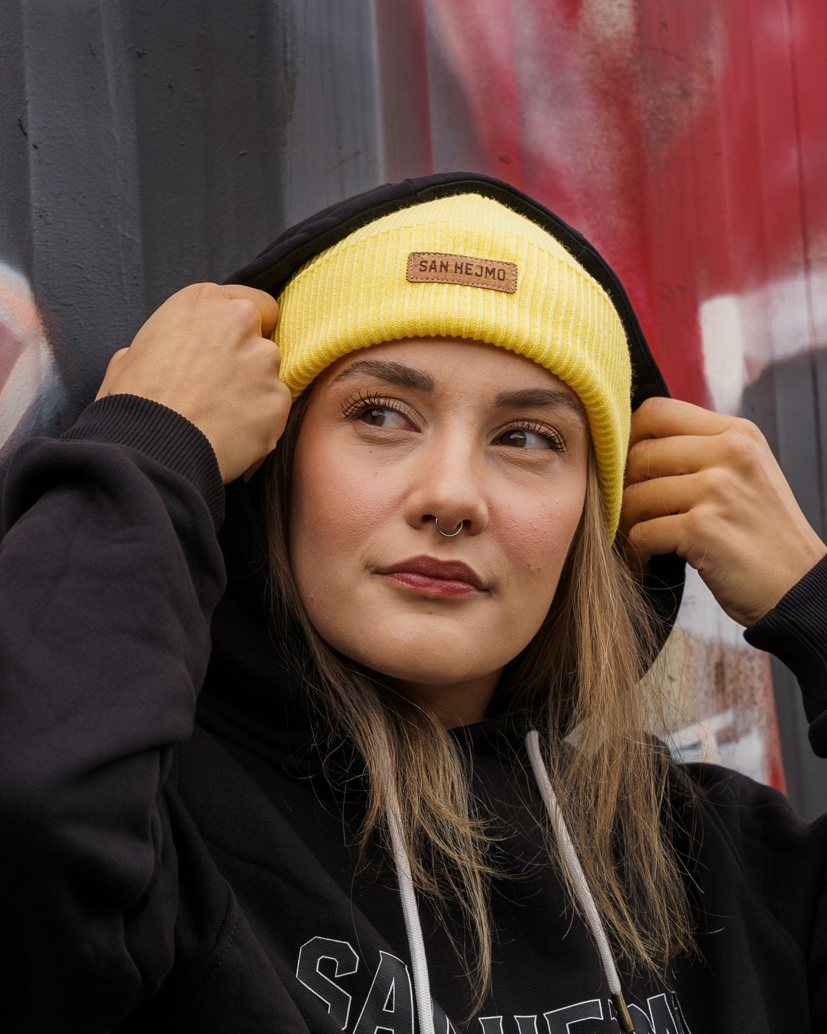 Eine Frau vor einem angesprühten Container, Sie trägt einen schwarzen San Hejmo Pullover und die gelbe Beanie aus der Basic Collection. Vorne ist ein braunes Tag aufgenäht auf welchem "San Hejmo" in dunkelbraun/schwarz gedruckt ist.