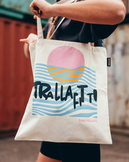 Ein naturbelassener beigefarbener Beutel mit der schwarzen Aufschrift „Trallafitti“. In einem künstlerischen Stil ist hinter diesen Worten ein Sonnenuntergang einer rosa Sonne am Meer zu erkennen. Die blauen Wellen sind als diverse Linien dargestellt.