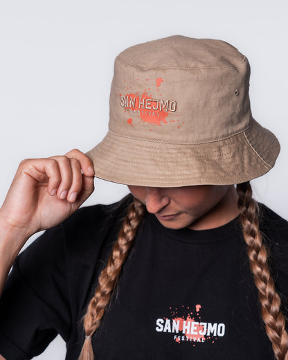 Ein wendbarer beigefarbener Bucket Hat mit der Aufschrift San Heimo ebenfalls in beige gestickt. Hinter der Aufschrift ist ein Farbklecks in orange abgedruckt.