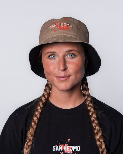 Der wendbare Bucket Hat mit der beigefarbenen Seite nach außen. Getragen wird er von einer jungen Frau. Der mittige beigefarbene Stick "San Hejmo" ist mit einem orangenen Farbklecks hinterlegt. 