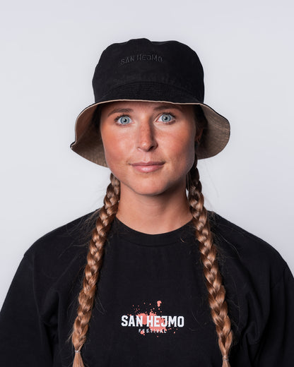 Der wendbare Bucket Hat mit der schwarzen Seite nach außen. Getragen wird er von einer jungen Frau. Der mittige schwarze Stick "San Hejmo" ist sichtbar.