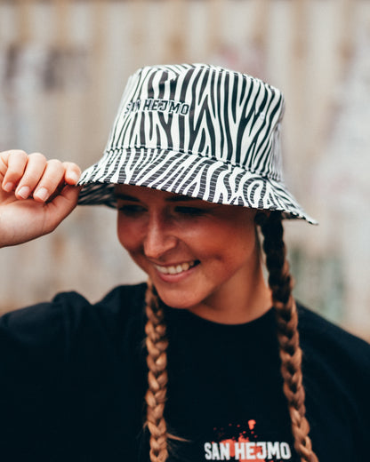Ein Bucket Hat mit Zebramuster. Mittig zu sehen, sind die Worte „San Hejmo“ in gestickter Form. Der Stick ist schwarz mit einem weißen, dünnen Rahmen. Getragen wird er von einer jungen Frau.