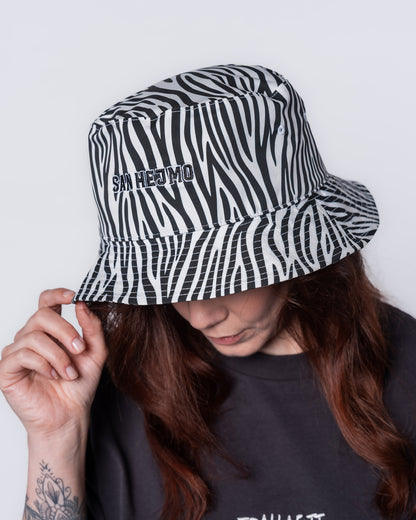 Ein Bucket Hat mit Zebramuster. Mittig zu sehen sind die Worte „San Hejmo“ in gestickter Form. Der Stick ist schwarz mit einem weißen, dünnen Rahmen. Getragen wird er von einer jungen Frau.