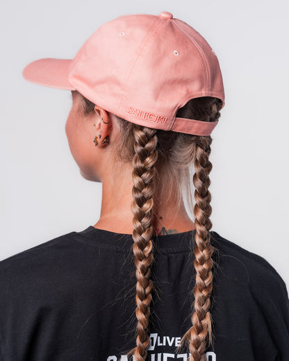 Eine Frau trägt das schwarze Line-up T-Shirt und die Apricot farbige Dad Cap. Sie steht mit dem Hinterkopf zur Kamera. Hinten links auf der Cap sind die Worte "San Hejmo" in Großbuchstaben gestickt.