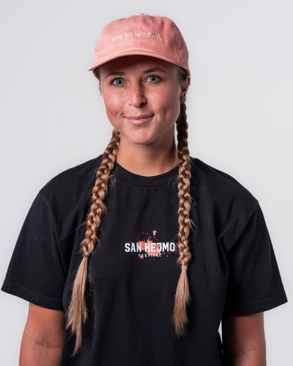 Eine Frau trägt das schwarze Line-up T-Shirt und die Apricot farbige Dad Cap. Sie steht gerade zur Kamera. Vorne mittig auf der Cap sind die Worte "Musik an, Welt aus" in weiß in Großbuchstaben gestickt.