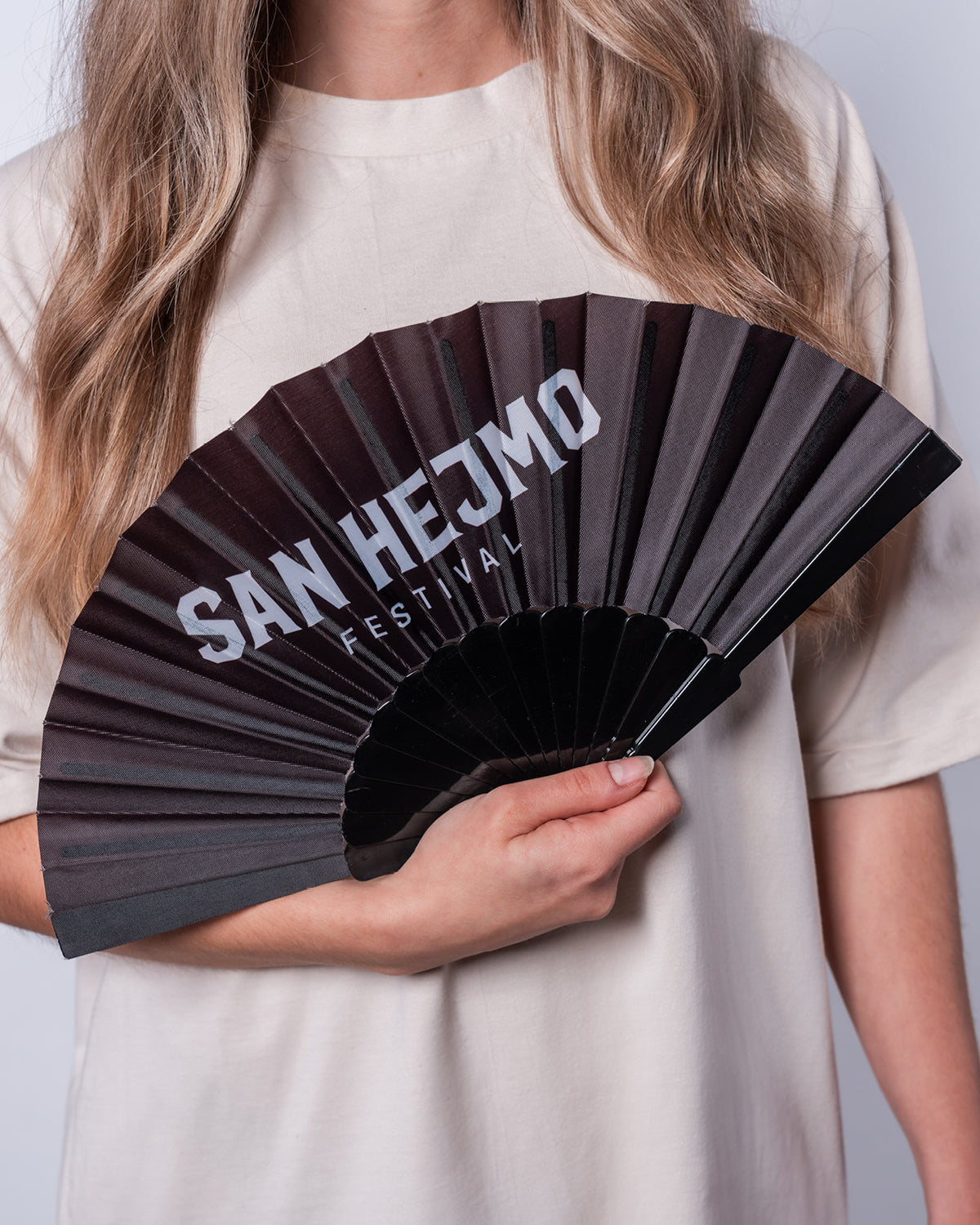 Ein schwarzer Fächer. Auf der oberen Stoffpartie sind die Worte „San Hejmo Festival“ in weiß gedruckt. Gehalten wird er von einer jungen Frau.