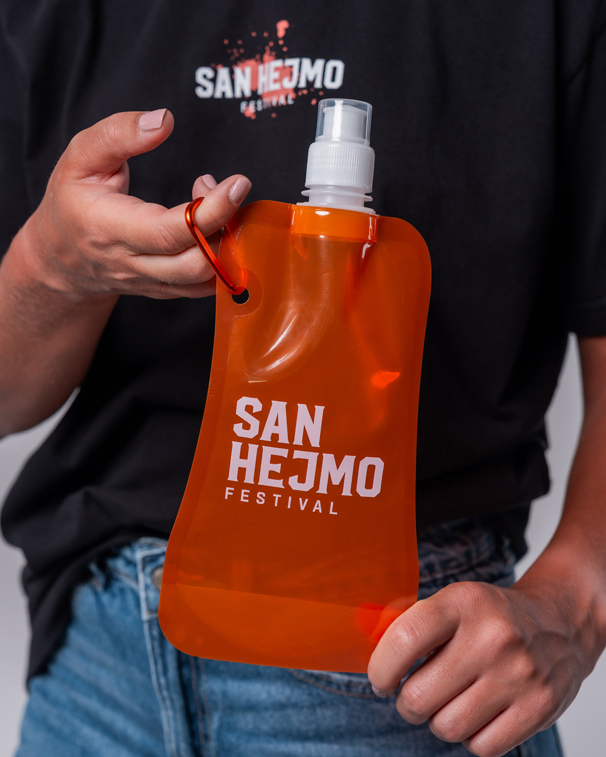 Eine orangene Kunststoffflasche mit einem 450ml Fassungsvermögen und Saugverschluss. Auf der Flasche ist der weiße Aufdruck "San Hejmo Festival" zu sehen. Die Flasche wird an einem orangenem Karabinerhaken festgehalten.