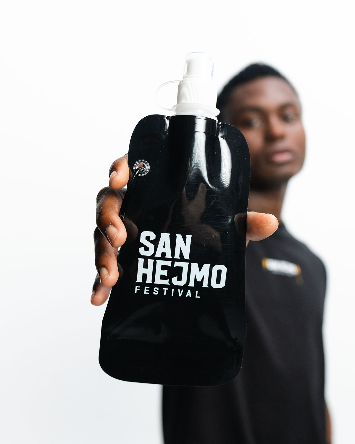 Eine schwarze Kunststoffflasche mit einem 450ml Fassungsvermögen und Saugverschluss. Auf der Flasche ist der weiße Aufdruck "San Hejmo Festival" zu sehen. Die Flasche wird von einem Mann in die Kamera gehalten. 