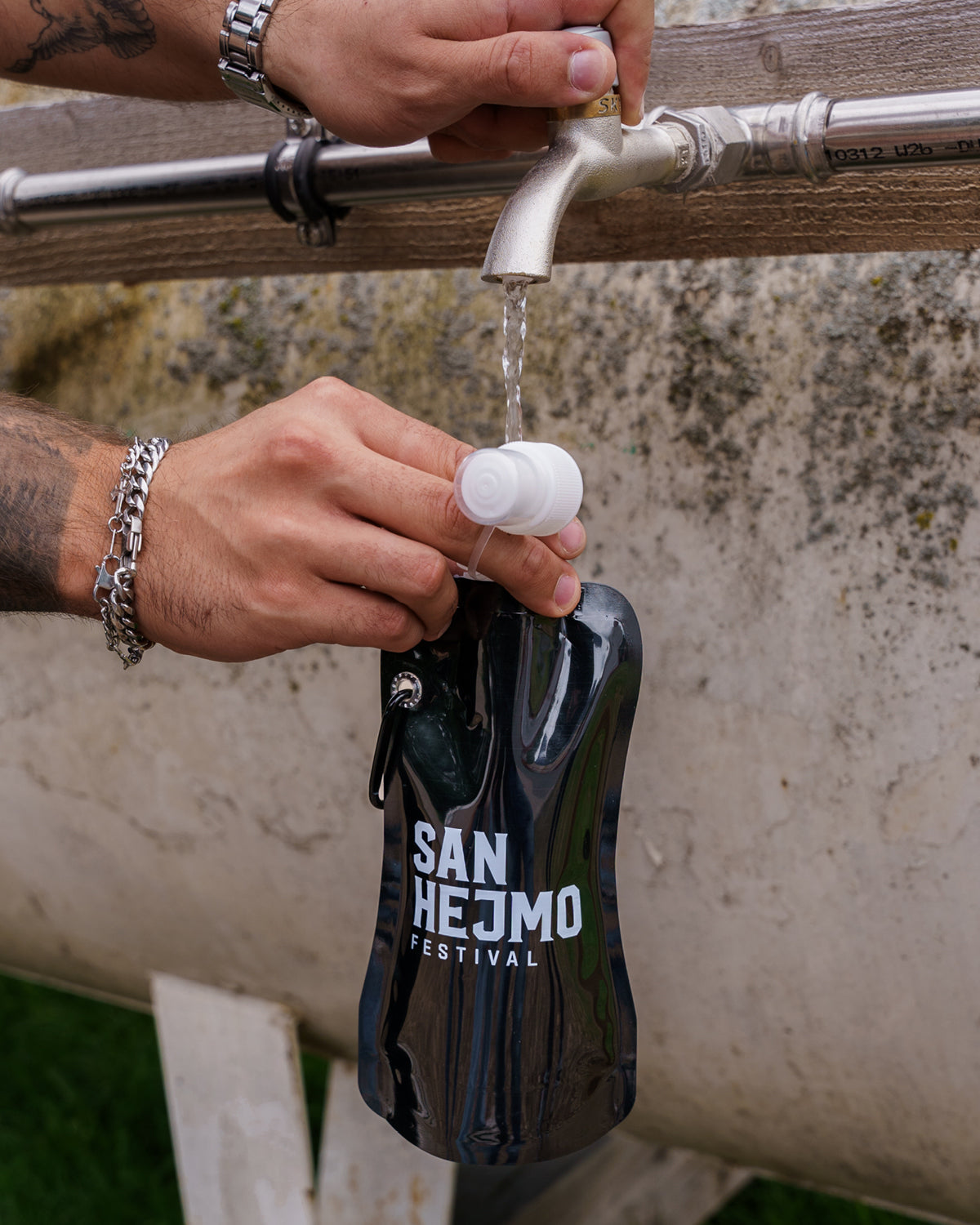 Eine schwarze Kunststoffflasche mit einem 450ml Fassungsvermögen und Saugverschluss. Auf der Flasche ist der weiße Aufdruck "San Hejmo Festival" zu sehen. Die Flasche wird von einem Mann unter eine Wasserstation gehalten und gefüllt.