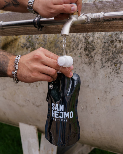 Eine schwarze Kunststoffflasche mit einem 450ml Fassungsvermögen und Saugverschluss. Auf der Flasche ist der weiße Aufdruck "San Hejmo Festival" zu sehen. Die Flasche wird von einem Mann unter eine Wasserstation gehalten und gefüllt.